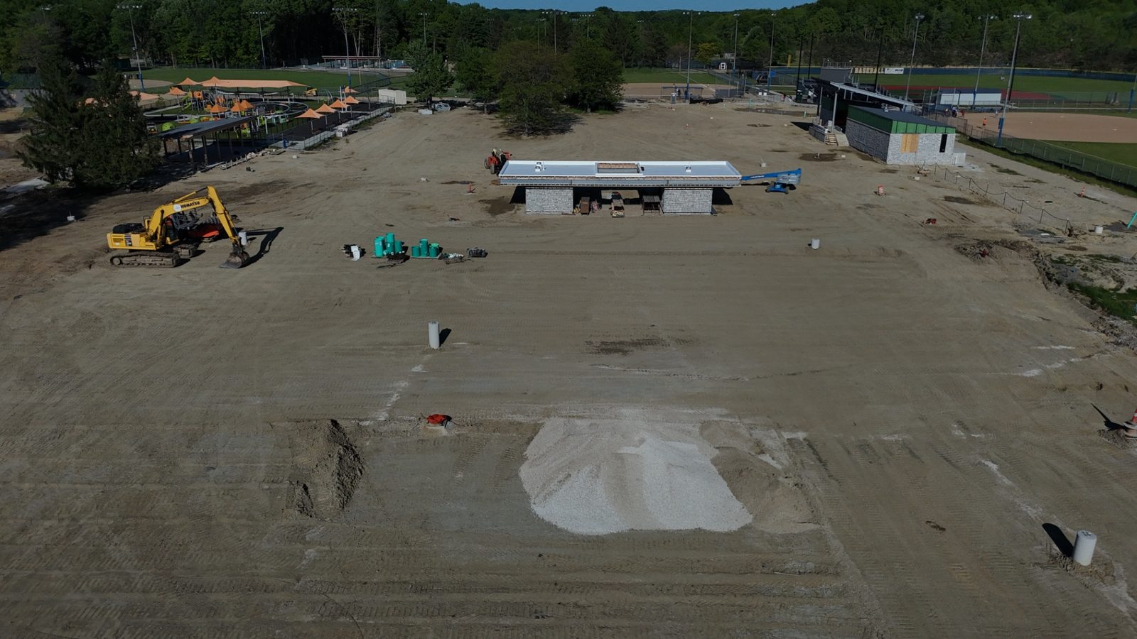 Aerial construction progress photo showing excavation work, exposed utility trenching, staged drainage pipe, and heavy equipment across the graded site — Stratum Aerial Media