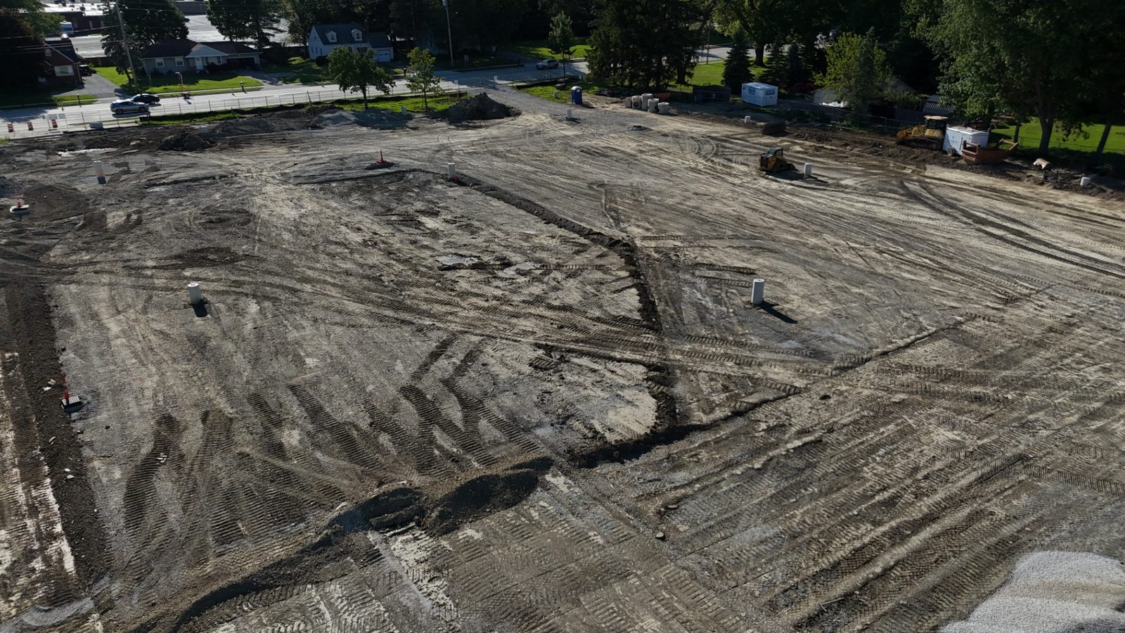 Aerial construction progress photo looking toward the street edge of the site, showing sitework, tire-tracked earthwork, equipment, and the surrounding neighborhood — Stratum Aerial Media