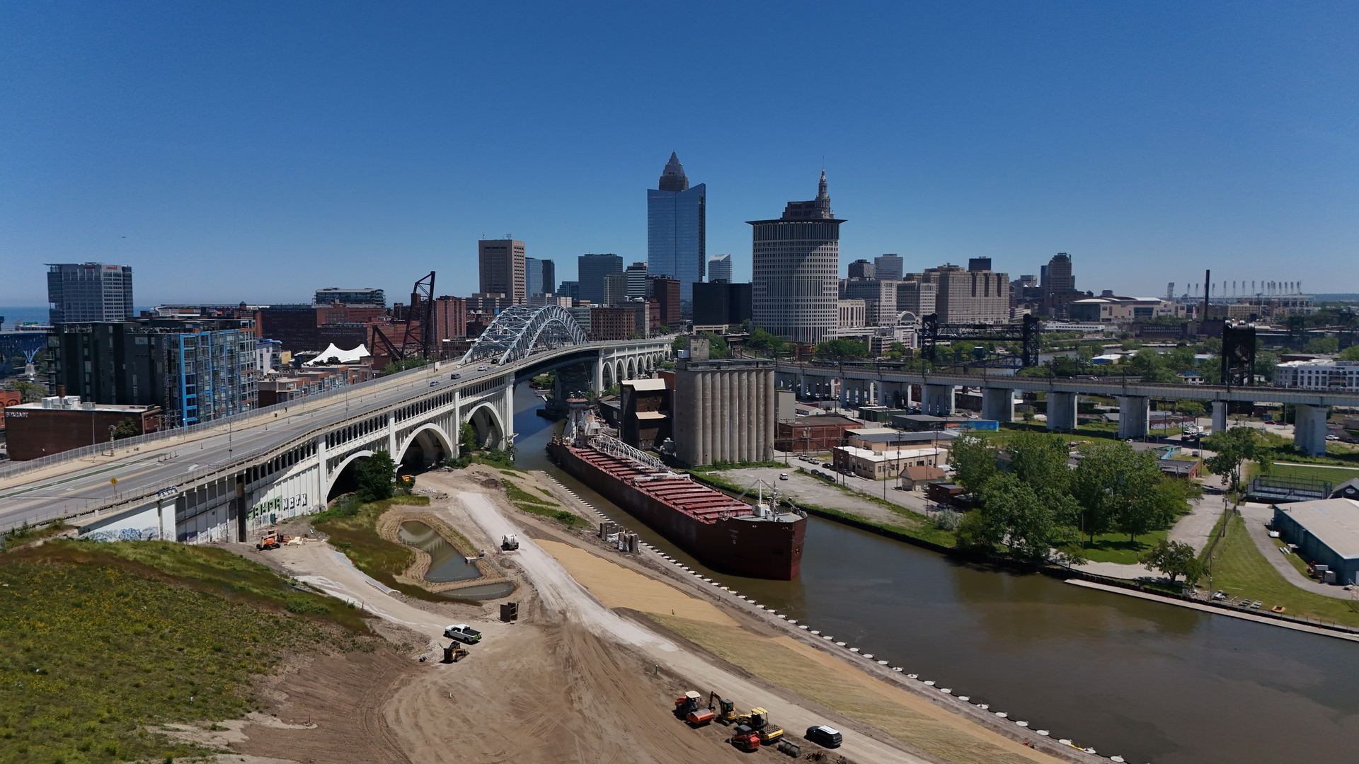 Aerial drone photograph of the Cleveland skyline behind the Detroit–Superior Bridge with a Great Lakes freighter on the Cuyahoga River and the Irishtown Bend construction site — Stratum Aerial Media